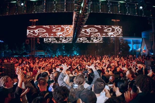 A panoramic crowd dancing in front of a large illuminated stage