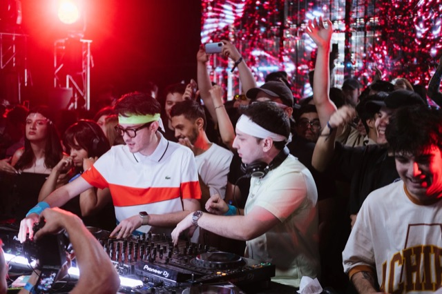 Two DJs playing inside a dancing crowd